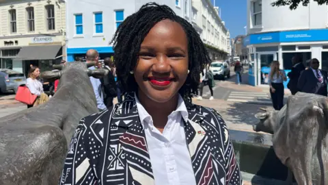Roseline Njogu is smiling as she stands outside in the sunshine in Jersey, with two statues of cows seen nearby.