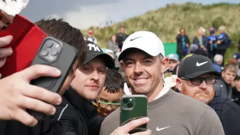 PA Rory McIlroy is smiling for a selfie with four or five young men. 

Rory is wearing a white nike hat and grey nike jumper. 

Behind him you can see the golf sand dunes and crowds of people gathering to see McIlroy. 

The sky is grey and the man closest to Rory is wearing a Yankee black cap and looking at his phone as he takes a selfie with Rory. 