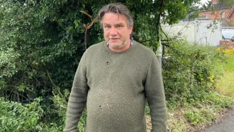 A man with short grey hair and wearing a green jumper stands at the entrance to a driveway in front of overgrown hedges.
