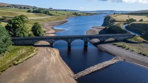 Andy Beck An aerial view of an old bridge emerging from a large body of water. A new modern bridge stands in the water just beyond it and joins a road curving up a hill. The water around the bridge is part of a larger body which stretches into the distance. The land to the left is covered in grass and trees.