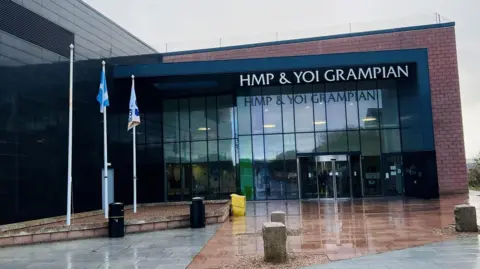 Brick and glass building with sign wording saying 'HMP & YOI Grampian', and flagpoles at the side.