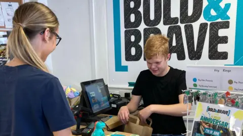 Two young people are standing by a cash register in a shop. A young man is packing items into a paper bag. Behind him the shop's name Bold & Brave is painted on the wall.