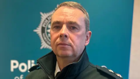 A police officer is looking down the lens of the camera. He has very short grey hair and is wearing 