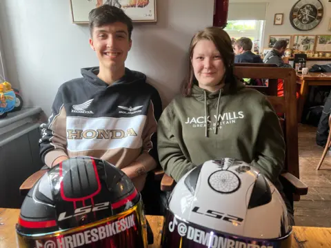 Harvey Mason and Izzie Childs are sitting at a pub table, smiling at the camera with their bike helmets on the table in front of them.