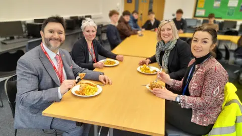 Gordano School Sadik Al-Hassan is sitting around a table with three women. They are eating lunch. They are all smiling at the camera.