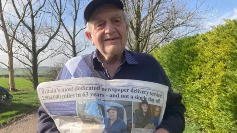 Alan Lawty pictured standing next to a hedge holding up a newspaper. He has grey hair and is wearing a blue jumper and cap.
