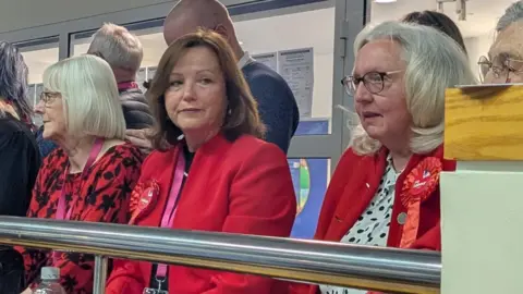Karen Clark is sitting and looking off the the right with a dejected look on her face. She has mid-length brown hair and is wearing a red jacket and red rosette. There are people sitting on either side of her.