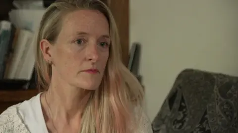 Catherine Milne is sitting on a sofa, in front of a bookcase. She has long blonde hair, with pink highlights.