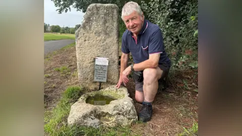 Ian Tomlinson kneels down in the grass beside two large stones; one is positioned upright and the other is basin-shaped, and filled with greenish coloured water. Ian is an older gentleman with short white hair, with a navy blue polo, brown shorts and black walking boots.