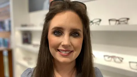 A woman with long brown hair and sunglasses perched on her head is smiling at the camera. She is wearing a denim coloured top and there are glasses in the background.