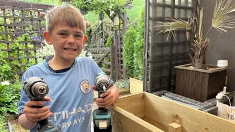 James who is holding two power tools and stood next to a wooden planter. He is wearing a light blue Manchester City shirt and has blonde hair, and smiles directly at the camera.