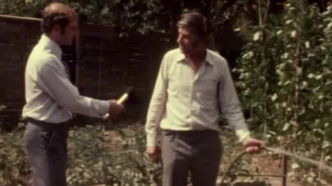 A man is holding a hosepipe full of bathwater. There are plants growing up canes. A reporter is standing near him and holding a microphone.