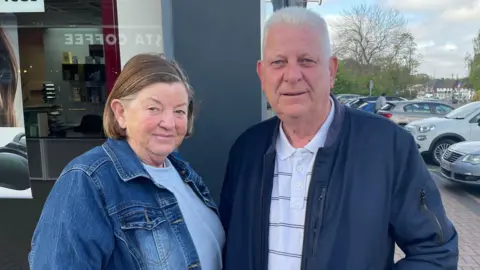 Daphne Bishop stands beside her husband William outside a row of shops in Aldridge town centre. She wears a denim jacket over a pale grey top. He wears a dark blue jacket over a white striped polo shirt.