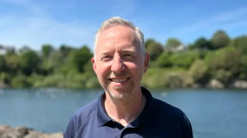 Mark Bowden smiling at the camera. He is stood in front of a reservoir with a bank with trees and grass behind it. 
