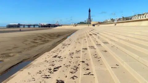 Steve Daniels/Geograph Steps leading on to Blackpool beach, with the tide out. Te tower and pier can be seen in the distance against a blue sky