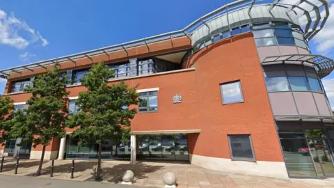 The exterior of Worcester Magistrates' Court set against blue skies on a sunny day. It's a modern building made of red brick, with curved glass at one end. Leafy trees stand on the street in front of the building.