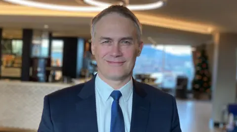 A man wearing a blue suit and blue tie smiles at the camera.