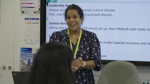 A teacher standing at the front of a classroom in front of a whiteboard. She is wearing a blue patterned dress and a yellow lanyard. The back of the heads of some pupils are in the foreground. 