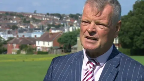 A TV still of Karl Love giving an interview in a park. He is wearing a blue pinstriped suit and a pink and white striped tie.