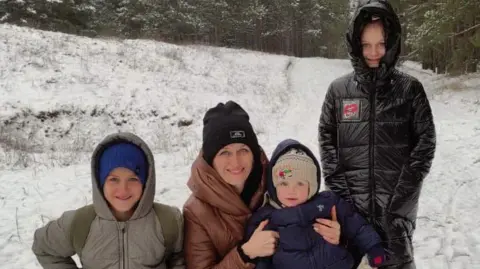 Fuzova Yuliia Fuzova is pictured kneeling down beside her three children in a snowy Ukraine. From left to right, the first child has a grey coat and blue hat, Fuzova is holding the second child in a navy puffer while she wears a brown coat and black hat. The third child is standing up and wears a black coat.