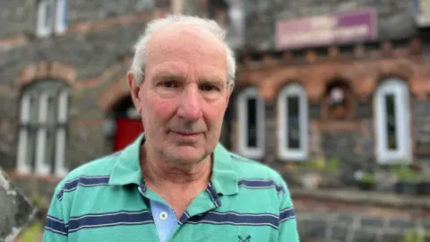 Richard Edwards, looking at the camera. He has thin grey hair, and is wearing a green and blue striped polo shirt. He is stood outside his guesthouse in Betws-y-Coed