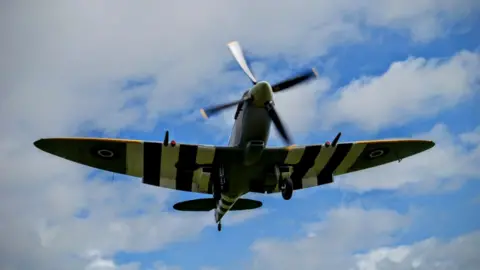  A spitfire which is light green and has black and white stripes on the wings flying against a blue sky with patches of white cloud. The propeller at the front of the plane is blurred as it is moving.