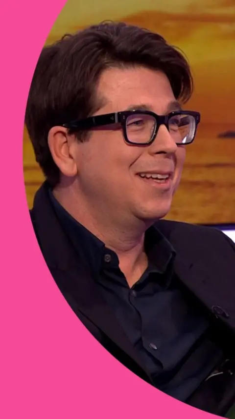 A man in a dark suit, with no tie, and glasses, sits smiling