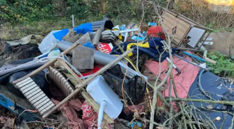 BBC A big pile of rubbish in a wooded area including chairs and other furniture.