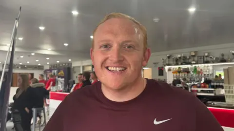 Louise Graham/BBC Louis Payne wearing a Nike top smiling at the camera. He has a row of trophies behind him.