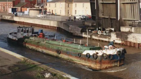 Contributed A long Dutch barge in disrepair with a green deck is sailing on a canal. There is a person on the barge at the end. It is sailing past a quayside where a number of cars are parked. 