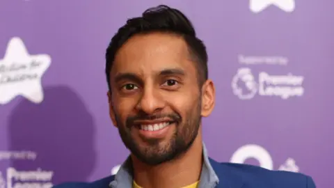 Bobby Seagull, a man with short black hair, wearing a yellow t-shirt, with a grey half-zipped jumper and a blue blazer, smiles for the camera while standing in front of a purple background.