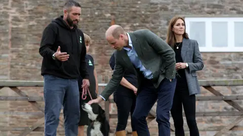 Reuters A man in a black hoodie and jeans talks as Prince William pets a black and white sheepdog he is holding on a lead. The prince is wearing a green blazer, blue shirt and navy trousers. Princess Catherine is wearing a white and black blazer, black top and trousers and her dark hair is down. A child with brown hair wearing black t-shirt and shorts can just be seen behind the man holding the dog. They are all outside, with a fence and a stone house behind them.