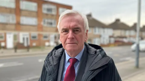 John McDonnell, a man with short grey hair wearing a dark coat, blue shirt and red tie