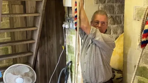 A man wearing a checked shirt pulls on a bell ringing rope. The red, white and blue striped rope hangs above him in the stone church tower.