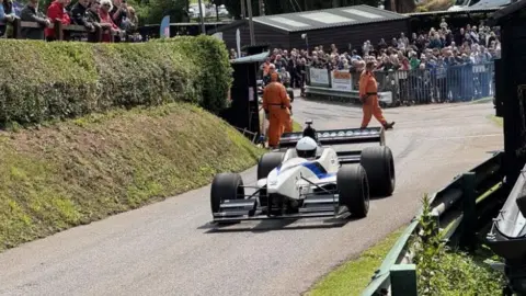 BBC A white Formula 1 style racing car on a tarmac track bordered by a grassy verge and embankment. Crowds stand behind a railing at the side of the track and lean over barriers at the side while crew in orange jumpsuits walk across it behind the vehicle.