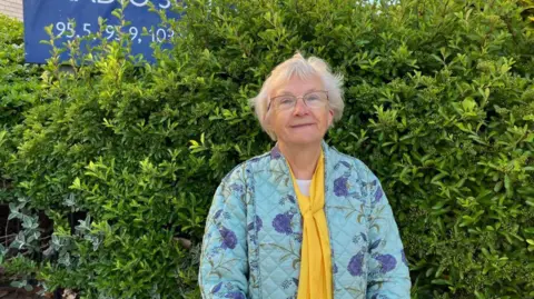 Vikki Irwin/BBC Inga Lockington is standing in front of the BBC Radio Suffolk building you can see a hedge to the right in the background and to the left there is a BBC Radio Suffolk sign. Inga is wearing patterned purple and teal padded coat with a yellow scarf. She is smiling at the camera. 