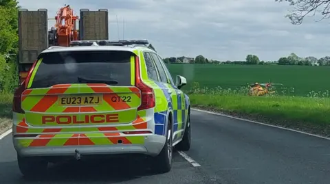 Essex Police A four-by-four police car parked on the single-carriageway road. A red and yellow air ambulance has landed on a large field to the right of the image.