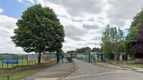 The entrance to a fenced school facility, with a wide paved driveway leading through a set of tall metal gates. On the left side of the entrance, there is an open grassy field bordered by a low brick wall and several signs attached to the fencing. A large tree with a full green canopy stands prominently on the left, casting some shade over the pavement. On the right side of the entrance, more trees and dense foliage grow behind another low wall, including one tree with dark purple leaves.
