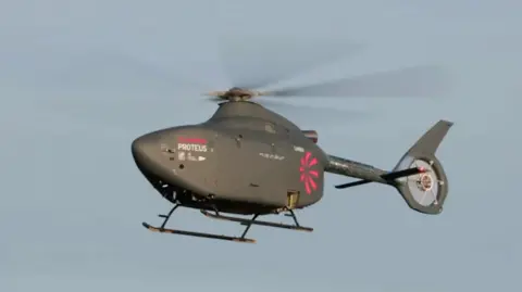 A helicopter without a window/cockpit flying in the blue sky. It is dark grey with the name Proteus on the front side in white and a pink logo of a rotor blade printed at the back