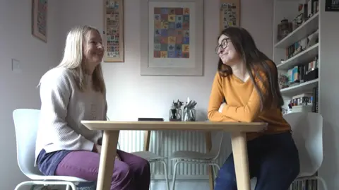 Katherine Bett and her mother Sue sitting opposite each other at a dining table. Katherine has long brown hair and is wearing glasses, a yellow jumper and blue jeans. Sue has mid-length blonde hair and is wearing a cream jumper and purple jeans. They are sitting on white plastic chairs. Behind them is a bookcase and there are pictures on the wall.