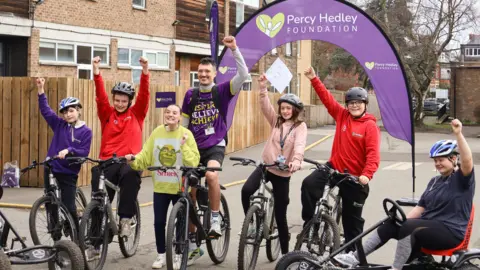 Garry Lee, a Special Support Assistant, stands under a purple archway with text that reads 'Percy Hedley Foundation'. He is wearing a purple t-shirt with the same text and riding a bike. He is joined by students with disabilities who are cheering. The students are riding trikes and bicycles.