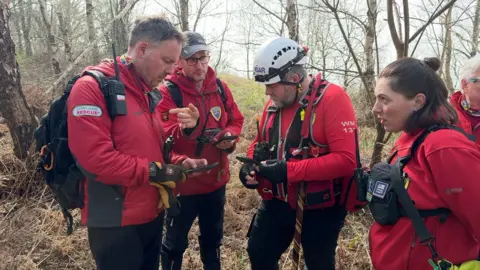 A group of volunteer rescuers in red coats and fleeces are stood in a huddle in woodland.