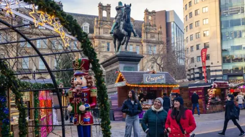 Christopher Heaney/Visit Leeds Wooden chalets dotted about a stone pavement Christmas decorated square. A statue of a man on a horse is in the background. Two women stand in the foreground, one in a black coat the other wearing a red coat.