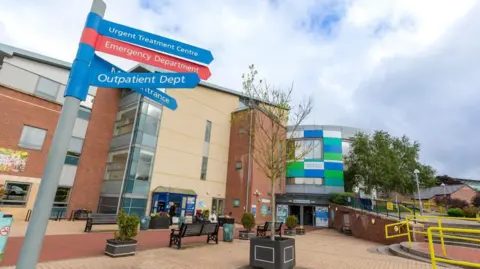 CDDFT The outside of the University of North Durham. It a brown and beige-coloured brick building. The entrance has green, white and blue rectangle panelling. There is a sign pointing patients to various departments. A small tree is in a plant pot in a seating area.
