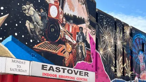 A colourful mural painted on a black background on a wall above the entrance to Eastover Indoor Shopping Centre. The mural includes a red train and two cowboys on horses as well as fireworks and an abstract illuminated fairground ride