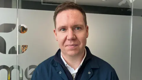 Jack Maclean/BBC Jonathan Mason, a man who is standing in front of a glass wall which has the Condimentum logo on it. He is looking directly at the camera and smiling and is wearing a white shirt with blue overalls over the top.