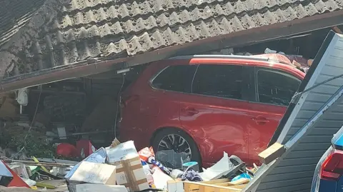 Cambridgeshire Constabulary Part of a red car can be seen crashed through a garage. The tiled roof of the building is on top of the car and a battered steel garage door can be seen off its hinges. There are boxes and other items that were inside the garage around the wheels of the car.