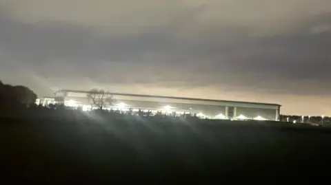 A brightly lit industrial unit, viewed from a field. There are trees between the photographer and the building, and the photograph has been taken at night.