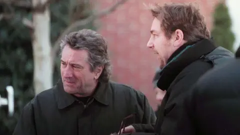 Getty Images Robert De Niro, with feathered hair and a dark jacket, stands outdoorsnext to another man, who is holding a pair of glasses and wearing a black jacket.