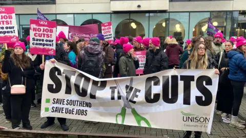 Dozens of people at a demonstration. In front of the picket line is a banner saying "Stop the Cuts". Some of the demonstrators are holding placards saying "No Job cuts". Many of them are wearing pink UCU branded hats.